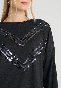 Nainen, jolla on tumma neulepaita, jossa on rintaosassa kaksi mustasta paljettikuvioista tehtyä viuhkakuviota ja kultaiset renkaakorvakorut.