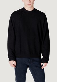 Maglione nero lavorato a maglia con un motivo a diamante texturizzato, dotato di scollatura a girocollo e polsini e orlo a coste, indossato con jeans scuri.