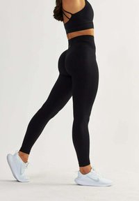 Leggings noirs à taille haute en tissu extensible, présentant une texture lisse, un design ajusté et des chaussures de sport blanches assorties.
