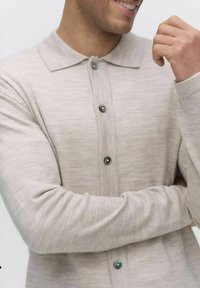 Ljust beige cardigan med krage, tillverkad av mjukt material. Har en knappfront med mörka knappar och figursydda ärmar.