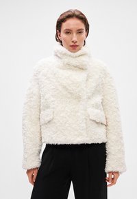 Biała kurtka z sztucznego futra z wysokim kołnierzem, frontowymi kieszeniami, fakturowaną powierzchnią i szerokimi rękawami, zestawiona z czarnymi spodniami.