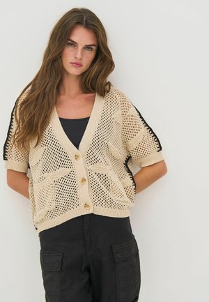 Mladá žena s dlhými hnedými vlasmi, oblečená v béžovom pletenom krátkornom kardigáne s gombíkmi cez čierne tričko a čierne pracovné nohavice.