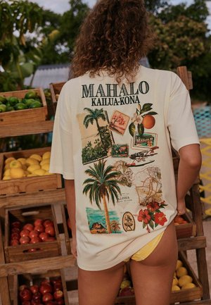Person iført en oversized beige T-shirt med tropisk Hawaii-tema print og tekst "MAHALO KAILUA-KONA", der står ved siden af trææsker med frugt.