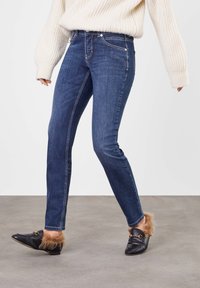 Dark blue slim-fit jeans with contrast stitching, worn with black loafers featuring fur accents. Featured alongside a cream knitted sweater.