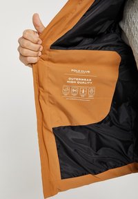 Pomarańczowa kurtka zewnętrzna z czarną podszewką, wyposażona w dużą kieszeń oraz metkę jakości z ikonami pielęgnacji i nazwą marki.