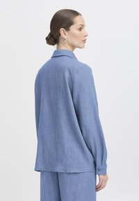 Blauwe langemouwige blouse met knoopsluiting en een relaxed fit, met subtiele verticale strepen en een zachte textuur. Achteraanzicht weergegeven.