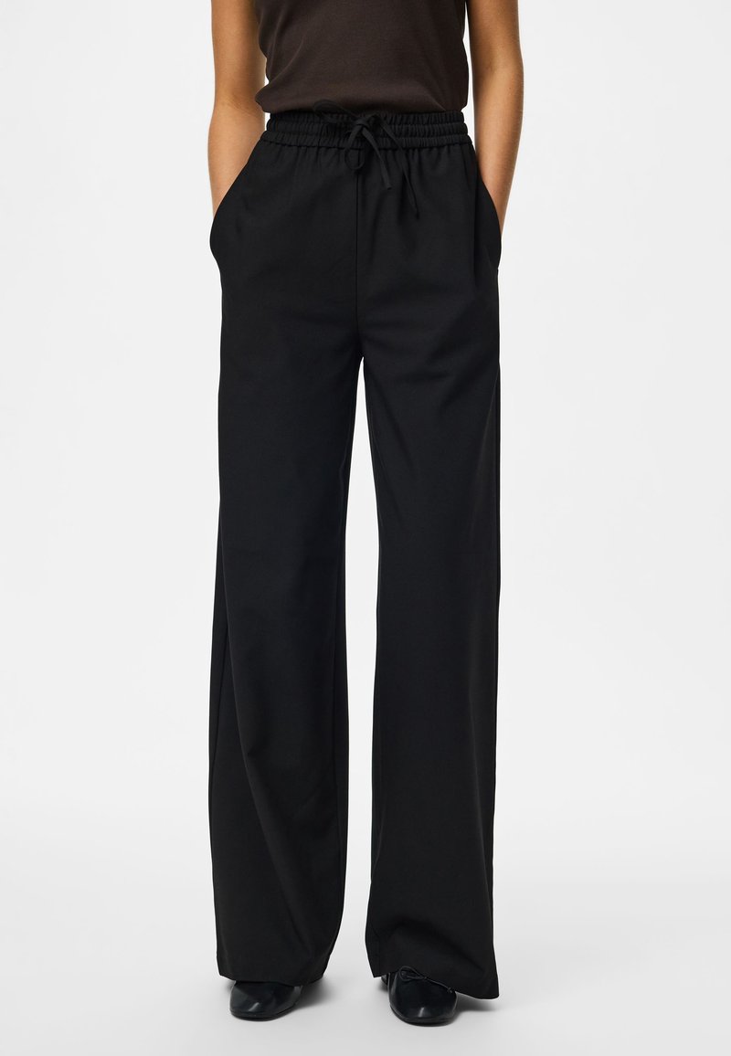 Persona que lleva pantalones holgados negros de pierna ancha con cintura elástica y cordón, manos en los bolsillos, combinados con zapatos negros y camiseta oscura.