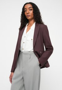 Blazer bordeaux con rever a V e dettagli del logo discreti, abbinato a una camicia bianca con bottoni e pantaloni a righe verticali grigio chiaro.