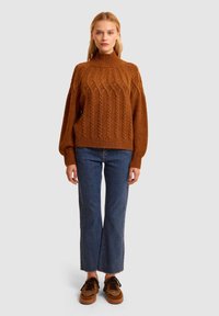 Maglione marrone lavorato a maglia con collo alto, caratterizzato da un motivo a treccia. Abbinato a jeans blu a gamba dritta e scarpe con stampa animale.