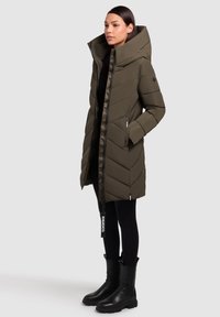 Pikk oliiviroheline puffer-jakk kõrge krae, lukuga ja chevron-õmblustega. Kombineeritud mustade retuuside ja massiivsete mustade saapadega.