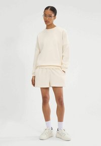 Crème sweatshirt met raglanmouwen, gecombineerd met crème shorts. Model draagt witte sneakers en subtiele witte sokken. Eenvoudig, casual ontwerp.