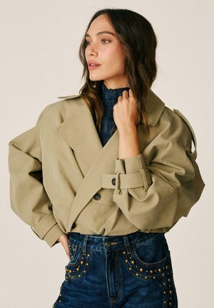 Mujer con una chaqueta beige oversize con correas con hebillas sobre un suéter azul oscuro y vaqueros de talle alto con tachuelas, mirando hacia la izquierda.