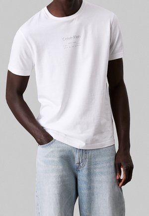 Mężczyzna ubrany w białą krótką koszulkę Calvin Klein i jasnoniebieskie dżinsy, z jedną ręką w kieszeni, na szarym tle.