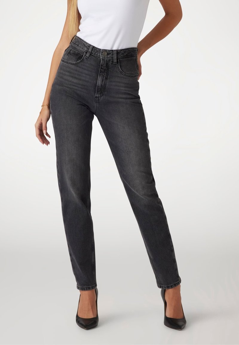 Femme portant un jean skinny noir taille haute, un top blanc sans manches et des talons noirs à bouts pointus, debout avec une main sur la hanche.