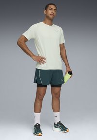 Lichtgroen sport T-shirt met korte mouwen, gecombineerd met donkergroene shorts met witte accenten. Zwarte hardloopschoenen en een felgroene waterfles.