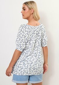 Blusa bianca a fantasia floreale con maniche a palloncino, caratterizzata da disegni floreali blu. Il retro presenta un collo arr gathered e una leggera lucentezza. Abbinata a pantaloni corti di denim.