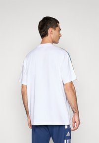 Hvit kortermet t-skjorte med rund hals, med blå og gule striper på skulderen. Tøyet ser glatt og lett ut.
