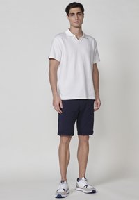 Vit strukturerad pikétröja för män med korta ärmar, i kombination med marinblå shorts och vita träningsskor med blå detaljer.