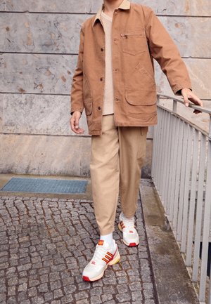 Giacca di tela marrone con tasche frontali, top beige in maglia e pantaloni beige. Scarpe da ginnastica bianche con accenti rossi e arancioni su ciottolato testurizzato.
