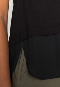 Top preto com design em camadas; a seção superior é feita de tecido suave, enquanto a seção inferior é ligeiramente transparente. Hemisfério arredondado, combinado com calças verde-azeitona.