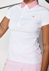 Bílé polo tričko s růžovým geometrickým vzorem na límci a bočních akcenty. Má krátké rukávy a malou nášivku na prsou.