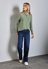 Maglietta verde chiaro a maniche lunghe con scollo a V abbinata a jeans a gamba larga blu scuro e sneaker bianche, su uno sfondo neutro.