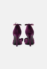 Sapatos de salto alto de veludo roxo, com traseira fechada, tira no tornozelo e salto agulha esguio. Superfície texturizada com acabamento suave.