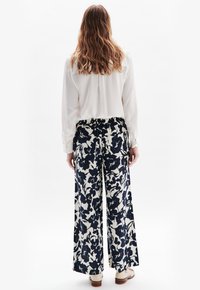 Vit blus med långa ärmar; kombinerad med vida byxor med ett marinblått blommönster mot en krämig bakgrund. Bruna skor synliga.