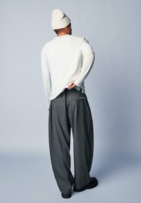 Weißes, geripptes Langarmshirt und graue, gestreifte weite Hosen mit einem Logo nahe der Gesäßtasche, getragen mit einer weißen Strickmütze.