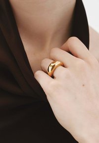 Anillo de metal dorado y redondeado en una mano, que muestra una superficie lisa y brillante con un diseño sutil. La mano reposa sobre una tela oscura.