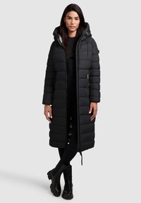 Manteau d'hiver long noir avec capuche, présentant un design matelassé, une fermeture éclair et deux poches latérales. Porté avec des vêtements ajustés noirs et des bottines.