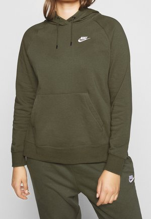 Person trägt olivgrünen Nike-Hoodie und passende Jogginghose, steht mit entspannter Haltung vor schlichtem Hintergrund.