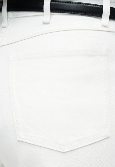 Κοντινή λήψη στην πίσω τσέπη λευκού τζιν με ορατά ράμματα και μαύρη δερμάτινη ζώνη περασμένη μέσα από τις θηλιές της ζώνης.