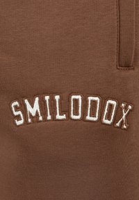 Brun sweatshirt med ribbet tekstur, der har "SMILODOX" broderet i hvid, buet skrift på den øverste frontsektion.
