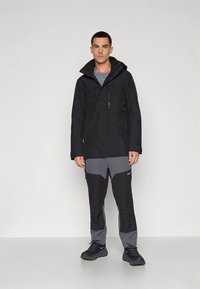 Melna ūdensnecaurlaidīga jaka ar augstu apkakli, divām priekšējām rāvējslēdzēm, kopā ar melnām un pelēkām biksēm ar sānu kabatām un pieguļošu dizainu.