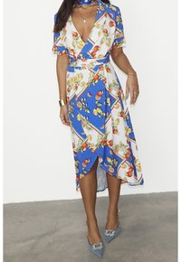 Robe portefeuille à fleurs dans des tons de bleu, blanc et vert. Manches courtes, décolleté en V, et ourlet au niveau du genou avec une coupe diagonale. Assortie à des talons bleus.