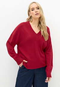 Roter V-Ausschnitt-Pullover aus weichem Strickmaterial, mit gerippten Bündchen und lässiger Passform. Kombiniert mit dunklen Jeans.