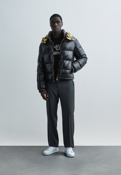 Pufferjakke i blank sort med udsmykket krave trykt med guld, båret over en sort skjorte og kombineret med grå bukser og hvide sneakers.