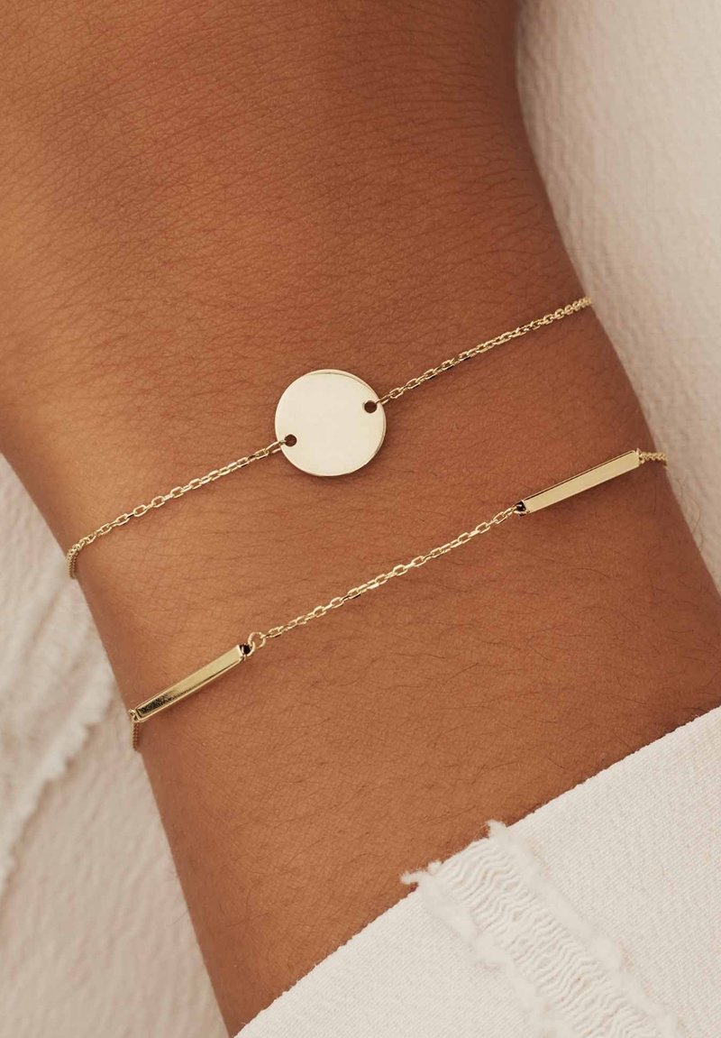 Gouden armband met een cirkelvormige hanger en een rechthoekige accent, beide verbonden door delicate kettingen, liggend op een huidkleurige pols.