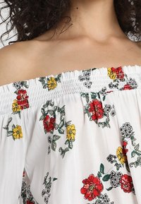 Off-shoulder vit topp med blommönster i röd, gul och grön. Har en smockad halsringning och lösa, flödande ärmar.