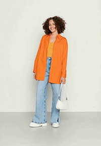 Camisa oversized laranja sobre uma blusa amarela, combinada com jeans flare azul claro e sapatos de plataforma brancos. Bolsa branca como acessório.