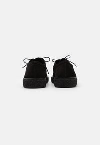 Scarpe nere in suede con suola nera texturizzata e lacci sottili, caratterizzate da una forma arrotondata e un design minimalista.