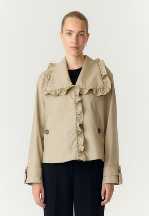 Femme portant une veste beige avec un col et un devant à volants, associée à un pantalon noir, debout devant un fond clair uni.