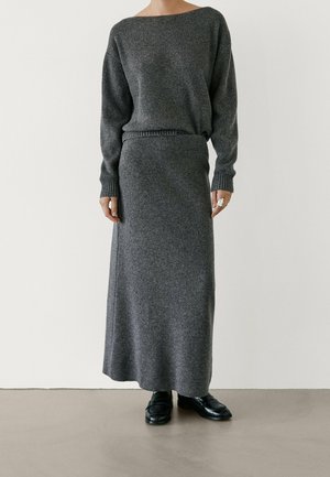 Femme portant une longue jupe en maille gris foncé et un pull côtelé assorti avec des mocassins noirs, debout contre un mur blanc uni.