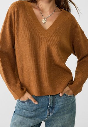 Kobieta ubrana w brązowy sweter z dzianiny z dekoltem w serek i długimi rękawami oraz niebieskie jeansy z denimu, ręce w kieszeniach i srebrny naszyjnik w kształcie serca.