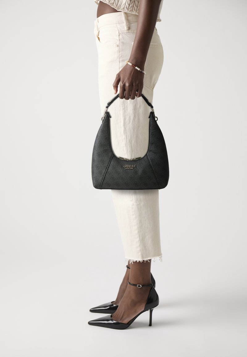 Personne tenant un sac à main GUESS noir, portant un jean crème, des talons hauts noirs à bride cheville et un pull court clair.