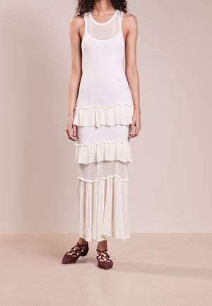 Femme portant une robe en maille blanche sans manches avec des volants superposés, associée à des ballerines pointues bordeaux ornées de perles et à des brides croisées.