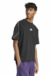 Junger Mann mit lockigem Haar, der ein schwarzes Adidas-T-Shirt mit weißen Paspeln und lila Hosen trägt, schaut nach links, die Hände in den Taschen.