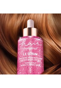 Flesje roze haarserum met een roségouden druppelaar, gelabeld "Hair Prodigieux Le Sérum," tegen een achtergrond van glad, glanzend bruin haar.