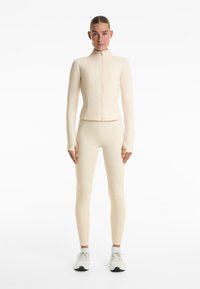 Giacca aderente a costine crema con colletto alto e zip completa, abbinata a leggings a costine coordinati. Modella che indossa scarpe da ginnastica bianche.